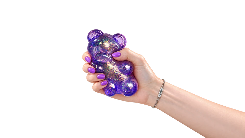 A hand squeezing a purple glitter bear SQUEEZU